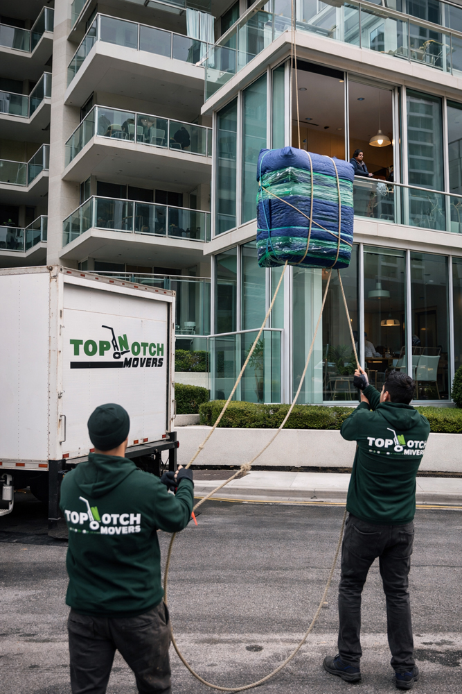 two male Top Notch Movers employees lowering a large household box via a rope and pulley system at a high-rise apartment